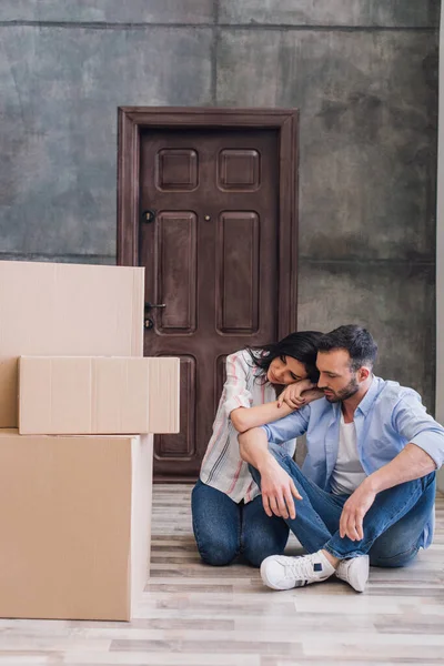 Empörte Frau lehnt sich mit gekreuzten Beinen an Mann in der Nähe von Boxen auf dem Boden im Zimmer — Stockfoto