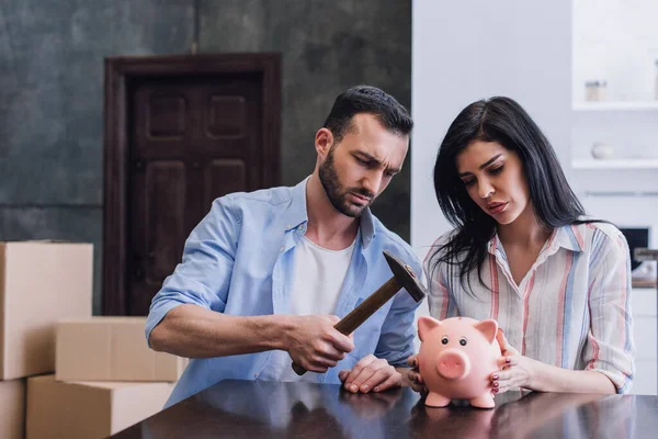 Woman with upset man holding hammer above piggy bank at table in room — Stock Photo