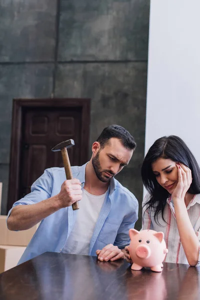 Mujer con hombre tenso sosteniendo martillo cerca de alcancía en la mesa en la habitación - foto de stock