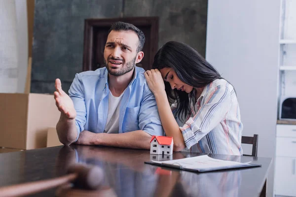 Selektiver Fokus einer aufgebrachten Frau, die sich an einen besorgten Mann am Tisch mit Hausmodell, Dokumenten und Hammer im Zimmer lehnt — Stockfoto