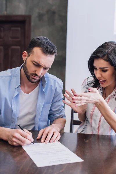 Gestresste Frau neben Mann schreibt Dokument an Tisch im Zimmer — Stockfoto