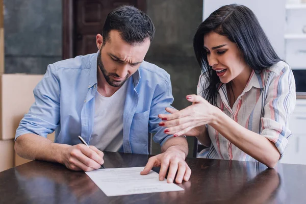 Stressed and worried woman near man writing in document at table in room — Stock Photo