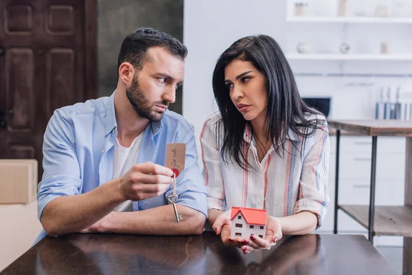 Upset bankrupts looking at each other and showing key with for sale lettering and house model at table in room — Stock Photo