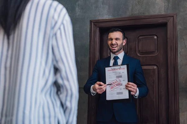 Angry collector pointing with pen at documents with foreclosure and final notice lettering in front of woman in room — Stock Photo