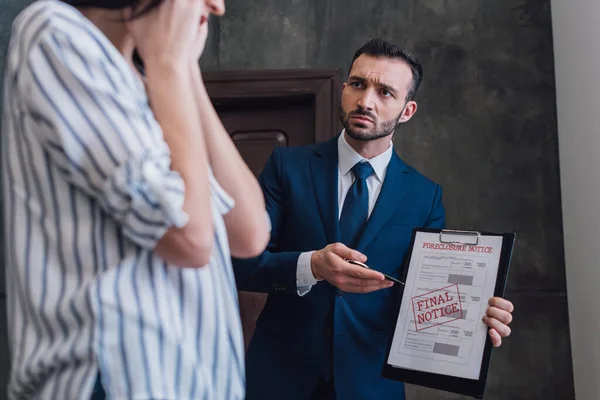 Collector pointing with pen at documents with foreclosure and final notice lettering and looking at woman in room — Stock Photo