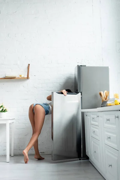 Vista laterale della ragazza sexy in pantaloncini frigo apertura in cucina — Foto stock