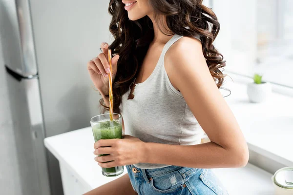 Vista recortada de una chica sonriente sosteniendo un vaso de batido en la encimera de la cocina - foto de stock