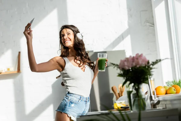 Selective focus of smiling girl in headphones holding smartphone and smoothie in kitchen — Stock Photo