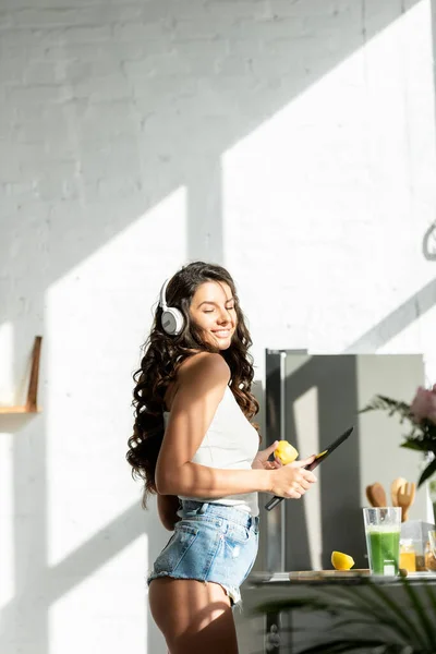 Side view of sexy woman in headphones smiling while holding piece of lemon and knife in kitchen — Stock Photo