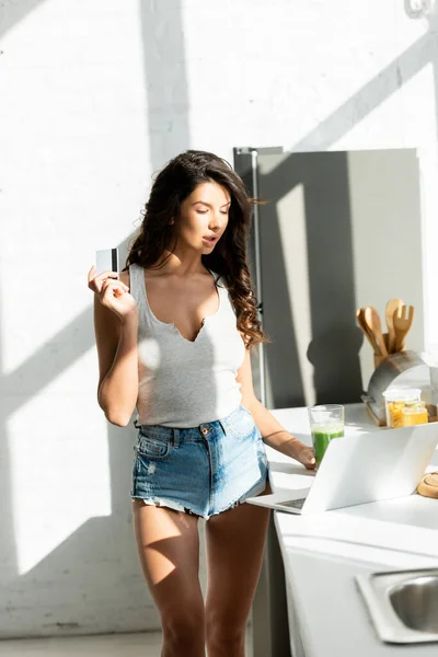 Hermosa mujer que sostiene la tarjeta de crédito mientras usa el ordenador portátil en la encimera de la cocina - foto de stock