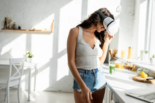 Focus selettivo della donna sorridente in cuffia realtà virtuale in piedi vicino al computer portatile in cucina — Foto stock