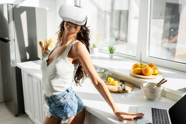 Chica sonriente en auriculares vr de pie cerca de la computadora portátil con tarjeta de crédito en la encimera de la cocina - foto de stock