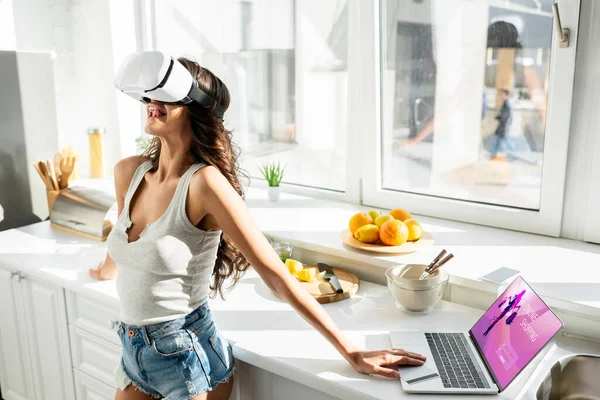 Girl in denim shorts using vr headset near laptop with online shopping website and credit card in kitchen — Stock Photo