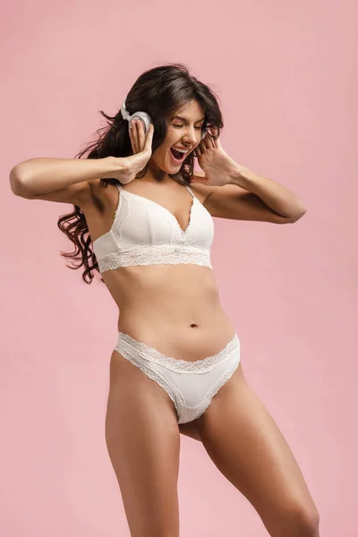 Femme excitée en lingerie toucher écouteurs et écouter de la musique isolée sur rose — Photo de stock