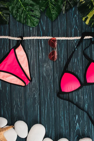 Top view of pink swimsuit near sunglasses, leaves and sea stones on dark wooden surface — Stock Photo