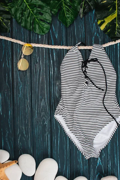 Vue du dessus du maillot de bain rayé près des lunettes de soleil, des feuilles et des pierres de mer sur fond de bois foncé — Photo de stock