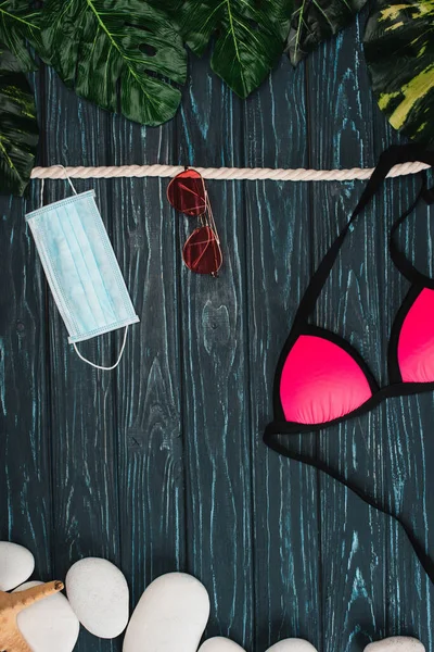 Top view of swimsuit, sunglasses and medical mask near leaves and pebbles on dark wooden background — Stock Photo