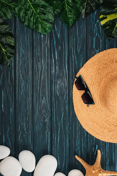 Vue de dessus du chapeau de paille et des lunettes de soleil près des feuilles et des pierres de mer sur fond de bois foncé — Photo de stock