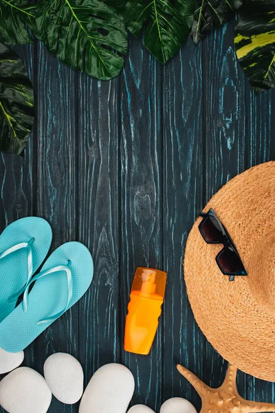 Vue du dessus de la crème solaire, des tongs près du chapeau de paille et des cailloux sur une surface en bois sombre — Photo de stock