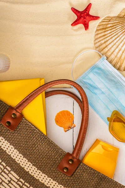 Vista dall'alto della maschera medica vicino alla protezione solare, borsa e libro su tavole di legno bianche vicino alle conchiglie sulla sabbia — Foto stock