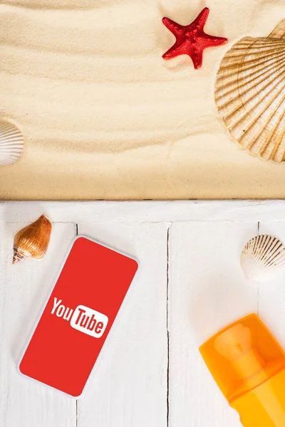 KYIV, UKRAINE - MARCH 25, 2020: Top view of smartphone with youtube app near sunscreen on white wooden planks and seashells with starfish on sand — Stock Photo
