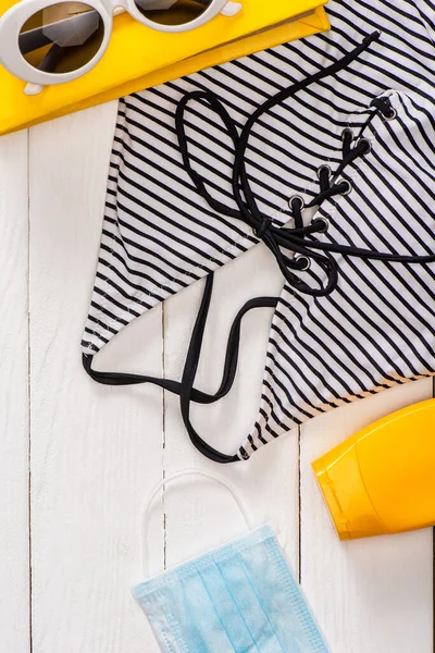 Vue du dessus de maillot de bain dépouillé, lunettes de soleil près du livre et masque médical sur fond de bois blanc — Photo de stock