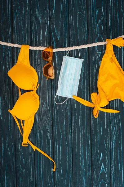 Vue du dessus du maillot de bain jaune près des lunettes de soleil et du masque médical sur corde sur une surface en bois sombre — Photo de stock