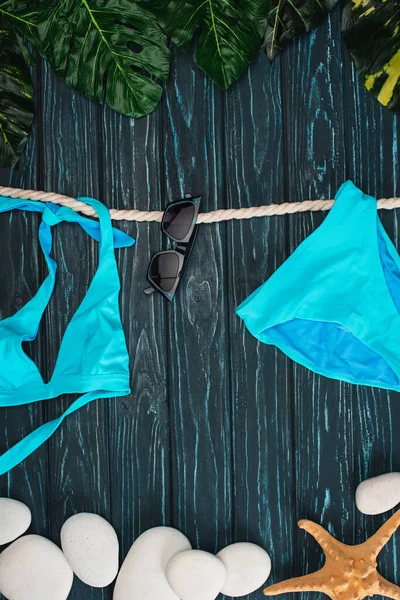 Draufsicht auf blauen Badeanzug in der Nähe von Sonnenbrille, Blättern und Meeressteinen auf dunkler Holzoberfläche — Stockfoto