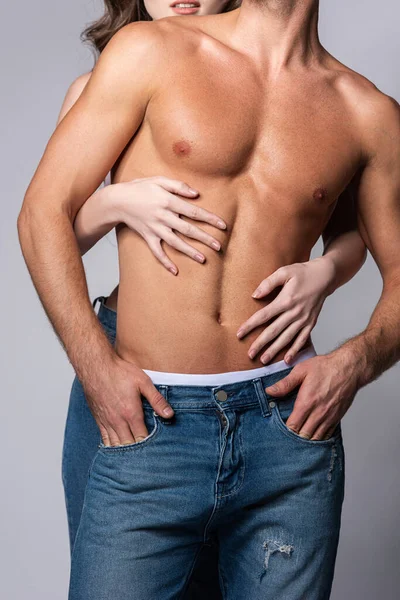 Cropped view of woman touching sexy and muscular boyfriend standing with hands in pockets isolated on grey — Stock Photo