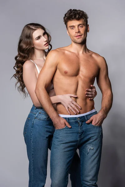 Frau berührt sexy und muskulösen Freund stehend mit Händen in Taschen auf grau — Stockfoto