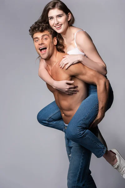 Excité et musclé homme piggybacking heureux copine sur gris — Photo de stock