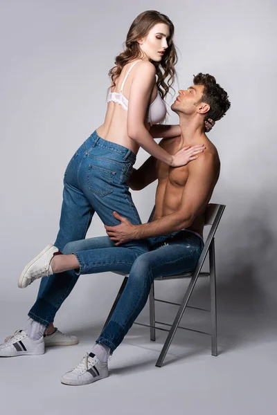 Seductive girl in bra touching muscular man sitting on chair on grey — Stock Photo