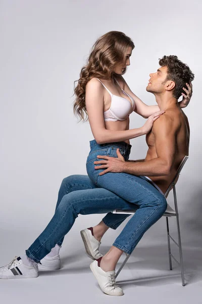 Vista lateral de chica seductora en sujetador sentado en el hombre musculoso en jeans en gris - foto de stock