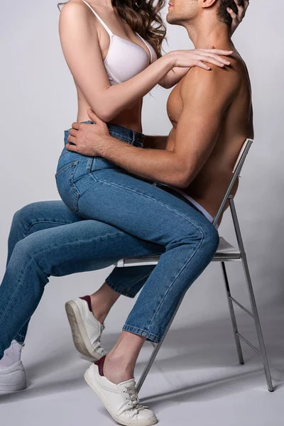 Cropped view of seductive girl in bra sitting on muscular man in jeans on grey — Stock Photo