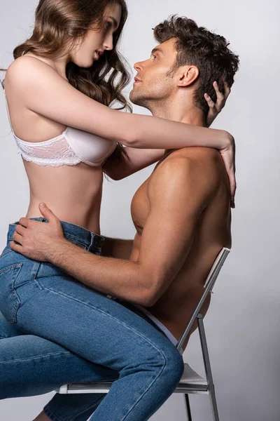 Profile of attractive girl in bra sitting on muscular man in jeans on grey — Stock Photo