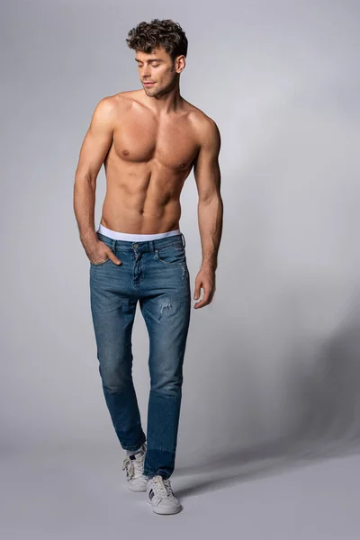 Muskulöser Mann in Jeans steht mit Hand in Tasche auf grau — Stockfoto