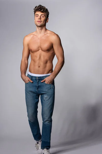 Homme musclé en jeans denim debout avec les mains dans les poches sur gris — Photo de stock