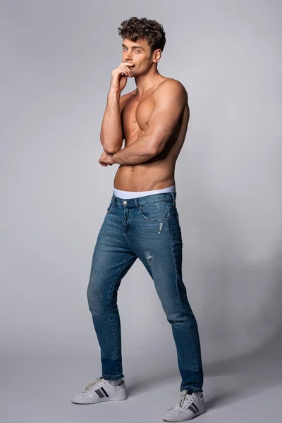 Мускулистый мужчина в джинсах в джинсах, смотрящий в камеру и трогающий лицо на сером — стоковое фото