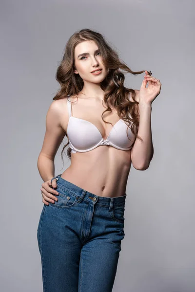 Seductive girl in bra and jeans touching hair on grey — Stock Photo