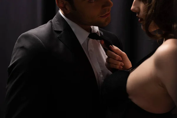 Cropped view of seductive woman pulling tie of man in suit on black — Stock Photo