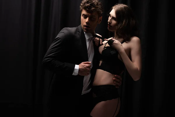 Sexy woman in underwear pulling tie of surprised man in suit on black — Stock Photo
