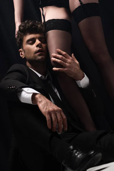 Selektiver Fokus eines gut aussehenden Mannes im Anzug, der das Bein einer sexy Frau in Strümpfen berührt und auf Schwarz sitzt — Stockfoto