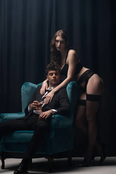 Jolie femme en sous-vêtements debout près de bel homme en costume assis dans un fauteuil et tenant un verre de whisky sur noir — Photo de stock