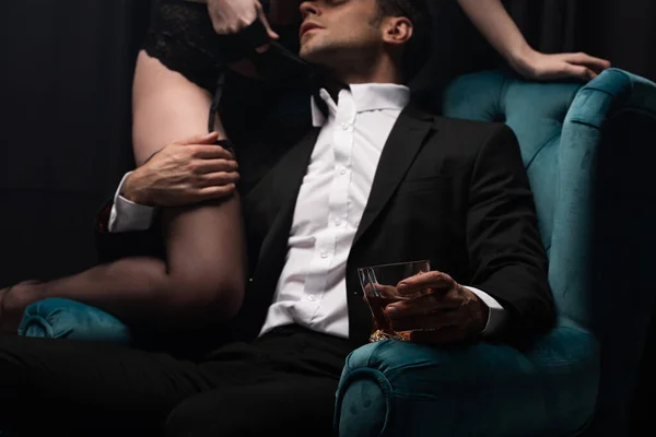 Обрезанный вид мужчины в костюме, сидящего в кресле, держащего стакан виски и трогательной соблазнительной женщины в нижнем белье на черном — стоковое фото