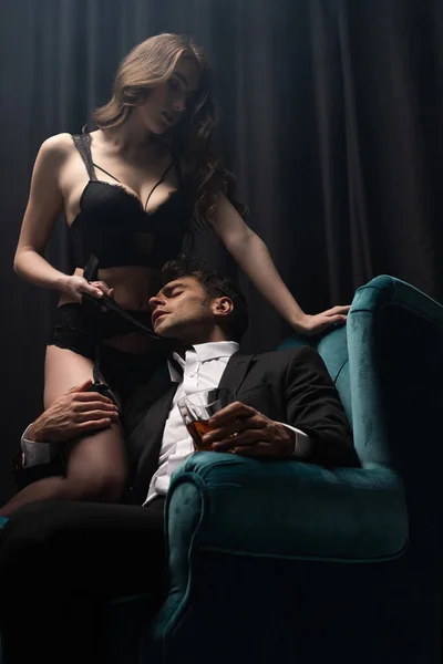 Homme en costume assis dans un fauteuil, tenant un verre de whisky et touchant femme séduisante en lingerie sur noir — Photo de stock