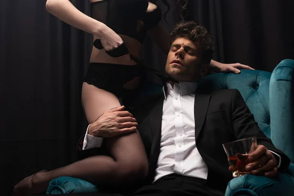 Uomo appassionato in giacca e cravatta seduto in poltrona, reggendo un bicchiere di whisky e toccando seducente donna in lingerie su nero — Foto stock