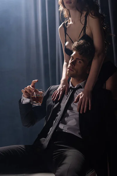 Donna seducente toccando bell'uomo seduto e tenendo un bicchiere di whisky sul nero — Foto stock