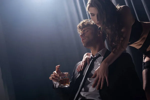 Seductora chica tocando guapo hombre en traje sentado y sosteniendo vaso de whisky en negro - foto de stock