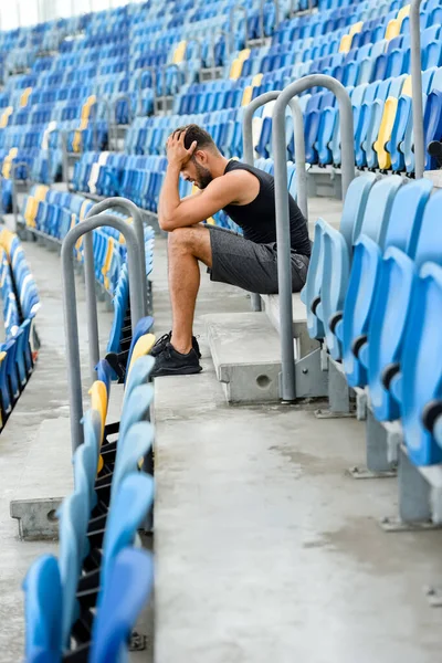Side view of young sportsman holding head while sitting on stairs near seats at stadium — Stock Photo