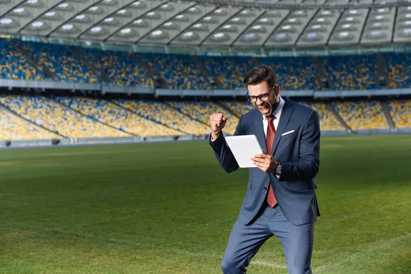 Giovane uomo d'affari in giacca e cravatta utilizzando tablet digitale e mostrando yeas gesto allo stadio, concetto di scommesse sportive — Foto stock